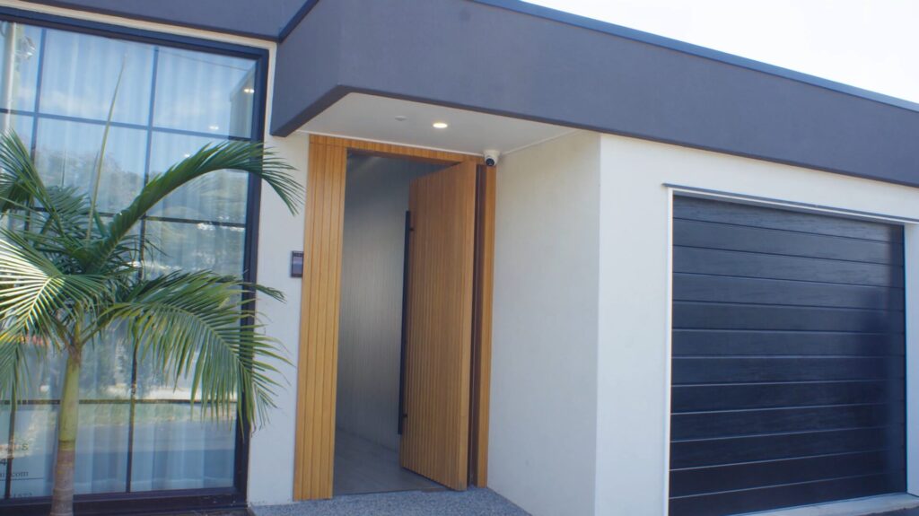 Modern house exterior with large glass window, black-framed garage door, light-colored walls, a wooden front door slightly open, and a small palm tree near the entrance.